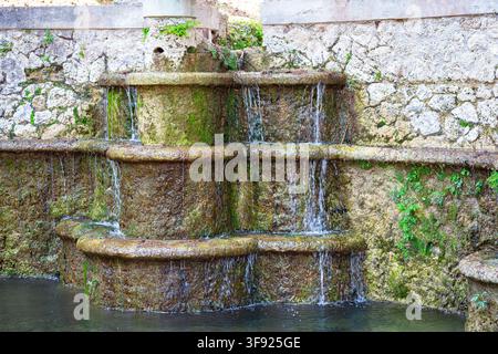 Fontaine en pierre à étages recouverte de mousse avec des cascades d'eau douces devant un mur de pierre rustique dans le jardin Banque D'Images