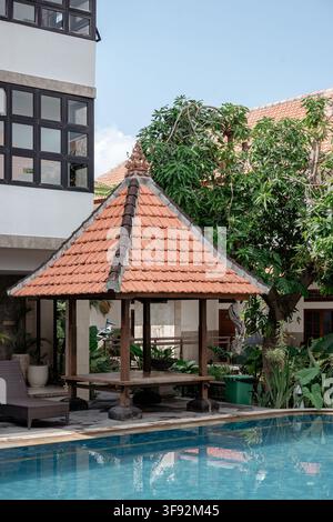 Une zone tranquille au bord de la piscine d'un complexe hôtelier avec un pavillon en bois traditionnel, une chaise longue, une eau bleue claire et un feuillage vert. Banque D'Images
