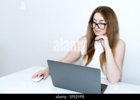 Une jeune femme portant des lunettes et un haut blanc assise à un bureau blanc minimaliste et propre, regardant pensivement l'écran d'un ordinateur portable gris Banque D'Images