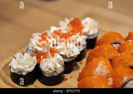 Vue rapprochée de rouleaux de sushi garnis de crème au fromage fouettée et de roe de poisson orange vif sur un plateau en bois, représentant une cuisine saine de qualité supérieure et v Banque D'Images