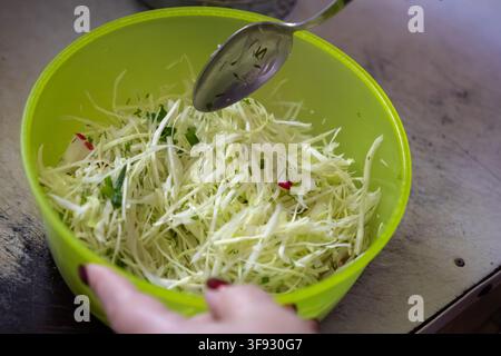 Une personne mélange du chou blanc tranché avec des herbes fraîches dans un bol en plastique vert citron pendant la cuisson, mettant en avant des habitudes alimentaires saines et des processus culinaires Banque D'Images