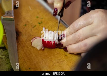 Prise de vue rapprochée de mains au vernis à ongles rouge coupant une radis croustillante en fines tranches sur une planche de bois rustique pour mettre en valeur les compétences culinaires et le bien-être Banque D'Images