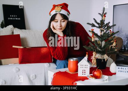 Jeune femme en bonnet du Père Noël tenant une tasse tout en étant assise sur un canapé près d’un petit sapin de Noël, boîte lumineuse HOLLY JOLLY. Réflexion sur l’année, résolutions du Nouvel An Banque D'Images
