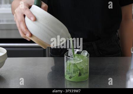 Une personne verse soigneusement du thé vert matcha d'une chawan en céramique dans un verre clair contenant des glaçons pour préparer un latte. Banque D'Images
