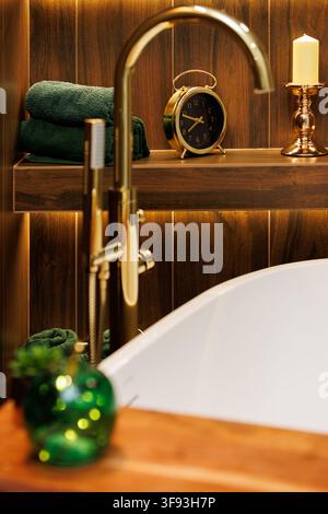 Un gros plan d'un robinet doré moderne à côté d'une baignoire blanche. Le fond présente une étagère en bois avec une horloge dorée, une bougie dans un porte-bougie, gr Banque D'Images