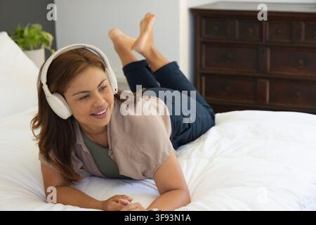 Femme allongée sur le lit dans la chambre, écoutant avec des écouteurs blancs, portant une chemise oversize beige et un jean Banque D'Images