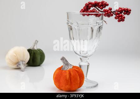 Un arrangement horizontal mettant en scène trois petits citrouilles en velours, une branche de baies rouges et un verre en cristal transparent. Cette composition élégante sur un fond blanc Banque D'Images