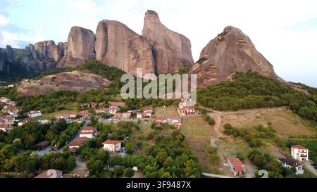 Vue aérienne époustouflante des immenses formations rocheuses de grès de Meteora dominant un village grec traditionnel niché parmi des arbres verts luxuriants. Banque D'Images