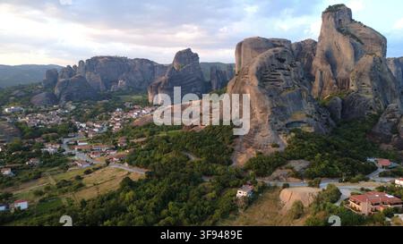 Pilier rocheux en grès dramatique dominant un village traditionnel niché dans une vallée verte, capturé depuis une perspective aérienne à l'heure dorée. Banque D'Images
