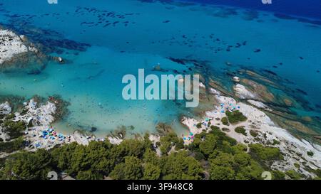 Vue aérienne époustouflante d'une crique de plage turquoise avec des formations rocheuses blanches, des nageurs et des parasols colorés entourés de pins. Banque D'Images