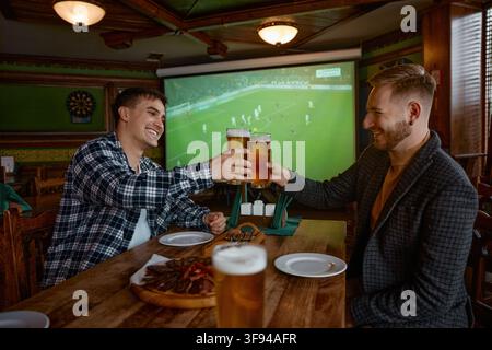 Deux hommes blancs trinquant des pintes dans un pub, des collègues célébrant un but en regardant le football sur un grand écran Banque D'Images