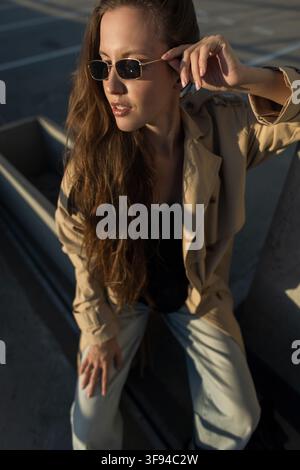 Jeune femme élégante aux cheveux longs, vêtue d'un trench-coat beige, d'un jean bleu et de petites lunettes de soleil, posant sur un parking urbain à l'heure dorée, béton Banque D'Images