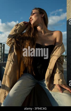 Jeune femme confiante aux cheveux longs et ondulés et portant des lunettes de soleil, vêtue d'un trench-coat beige lâchement porté par-dessus un haut noir et un jean clair, est assise sur un banc en béton Banque D'Images