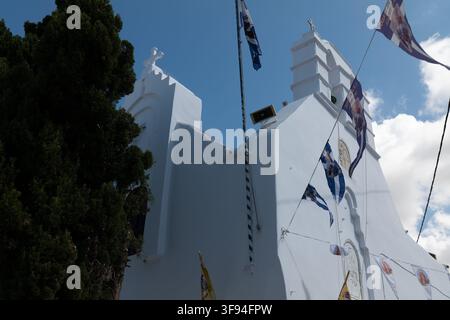 Chapelle blanche avec des drapeaux grecs et des guirlandes à côté d'un arbre sous un ciel bleu, Mykonos, Grèce. Banque D'Images