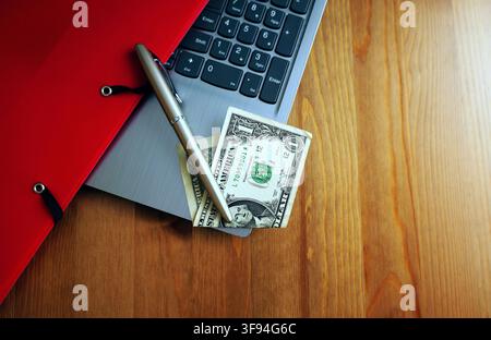 Clavier d'ordinateur portable, dossier rouge, stylo et billets de banque en dollars américains posés sur un bureau en bois, symbolisant la planification financière, l'intégration technologique et les affaires m Banque D'Images