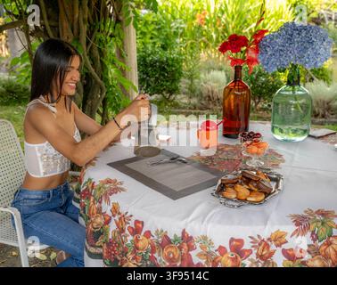 Jeune femme souriante versant du café d'une moka à une table de jardin recouverte d'une nappe fleurie Banque D'Images