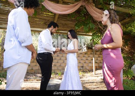 Couple échangeant les anneaux sous une pergola drapée de tissu rose dans le jardin, la mariée portant une robe de dentelle blanche Banque D'Images