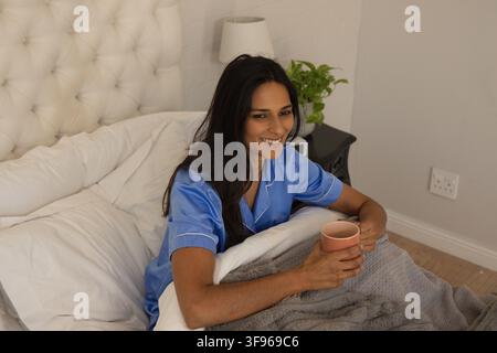 Femme assise dans un lit tenant une tasse en céramique rose, portant un haut de pyjama en satin bleu, souriante Banque D'Images