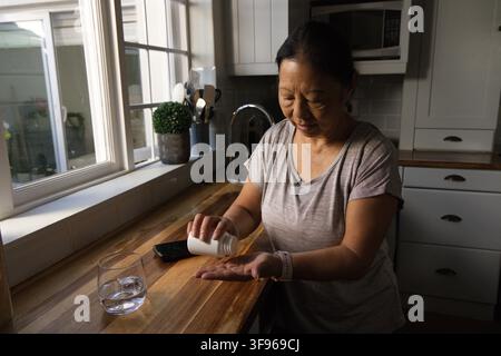 Sénior asiatique versant des pilules d'une bouteille blanche dans la paume à un comptoir en bois avec téléphone, eau Banque D'Images