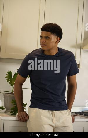 Homme afro-américain début de la vingtaine appuyé sur un comptoir dans la cuisine portant un T-shirt bleu marine, plante et poêle Banque D'Images
