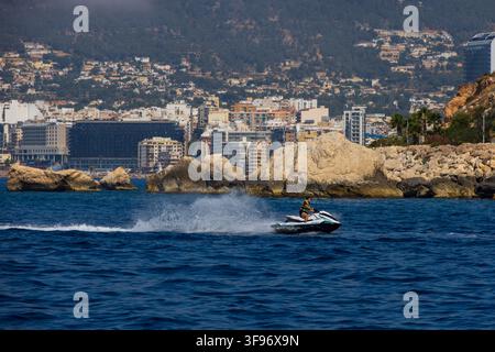 Calp, Espagne - 11 juillet 2027 : Vue d'une personne pilotant un jet-ski sur la mer Méditerranée avec la ville côtière de Calp et des falaises rocheuses en arrière-plan Banque D'Images