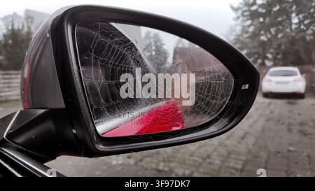 Prise de vue rapprochée détaillée du rétroviseur latéral d'une voiture recouvert d'une toile d'araignée givrée et complexe, capturant un reflet unique d'un matin d'automne et créant une Banque D'Images