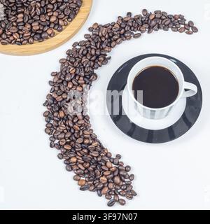 Vue de dessus du café noir dans une tasse rayée à côté d'une traînée coulant de grains de café torréfiés et d'un plateau en bois sur fond blanc avec un espace de copie utile. Banque D'Images