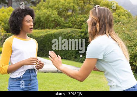 Différentes amies féminines debout et en train de discuter sur une pelouse dans un parc avec des lunettes de soleil posées sur la tête Banque D'Images