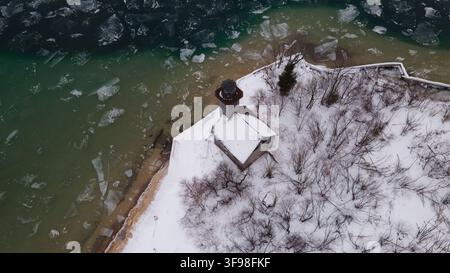 Vue aérienne du phare de Grand Island entouré d'arbres enneigés et des eaux gelées du lac Supérieur à Munising, Michigan, États-Unis. Banque D'Images