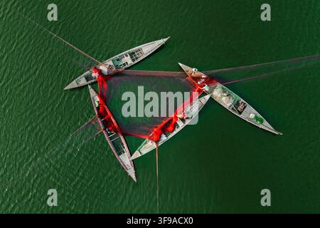 Vue aérienne de trois bateaux en bois disposés en triangle, reliés par de grands filets de pêche rouges, avec des pêcheurs travaillant sur l'eau calme et verte de la rivière Banque D'Images