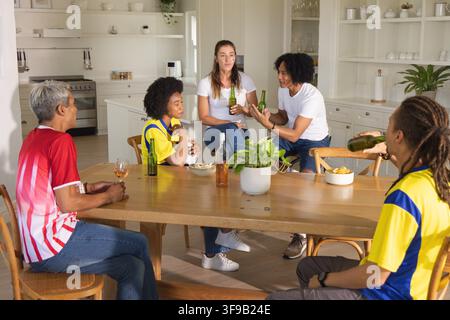 Cinq amis dont un homme âgé portant des maillots assis à la table de la cuisine à la maison, tenant une bière verte Banque D'Images