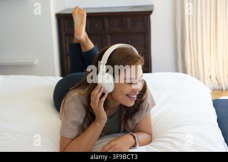 Femme africaine adulte allongée dans un lit écoutant avec un casque blanc, touchant l'oreillette, souriante, portant une montre Banque D'Images