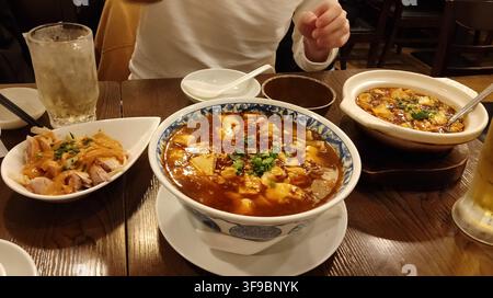 Un gros plan d'un ragoût authentique de tofu mapo servi dans un grand bol en céramique et un pot en terre cuite accompagné d'une entrée au poulet sur une table en bois Banque D'Images