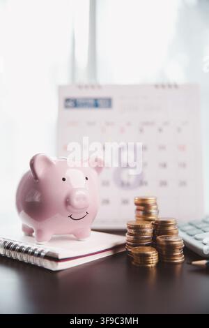 Cochon de banque rose sur un cahier à côté de pièces empilées, calculatrice et calendrier flou, symbolisant les économies mensuelles, le budget familial, le plan financier Banque D'Images
