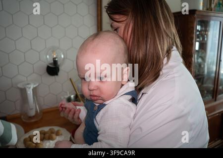 Mère tient son fils tout petit près d'elle tout en soutenant doucement sa main lors d'un moment familial à la maison. Maternité, soin maternel, garde d'enfants, affection, protection Banque D'Images
