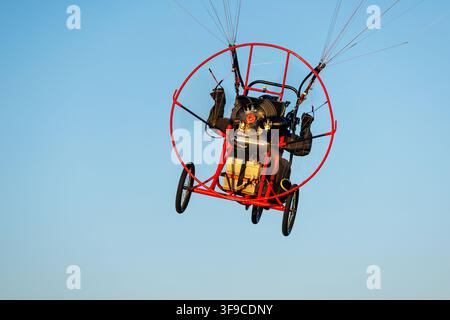 Un deltaplane motorisé avec un cadre métallique rouge vif et des roues planant haut dans le ciel bleu clair, capturé depuis un angle bas pendant une journée ensoleillée Banque D'Images