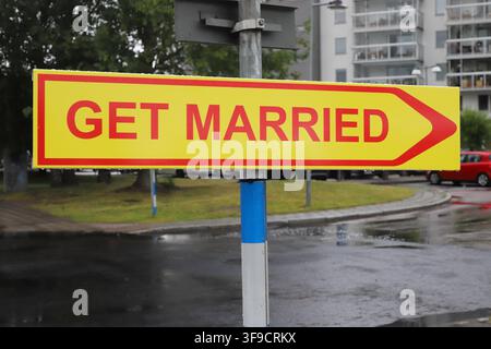 Grossissement d'un panneau jaune portant l'inscription "GET MARRIED" en lettres rouges avec une flèche pointant vers la droite, monté sur un poteau le long d'une rue dans un quartier résidentiel. Banque D'Images