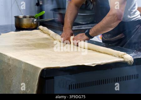 Boulanger habile roulant une plaque de pâte en forme de bûche sur un grand comptoir dans la cuisine. Artisanat traditionnel de fabrication de pain avec les mains façonnant une pâte fraîche dans Banque D'Images