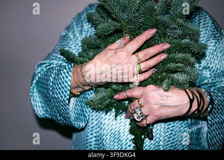 Une femme âgée tient une branche d'épicéa fraîche près de ses mains décorées, ornées de bagues et de bracelets, portant un chemisier à motifs bleus et blancs Banque D'Images