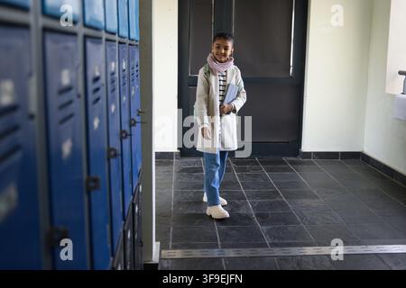 Étudiant afro-américain debout à l'école, vêtu d'un manteau et d'une écharpe rose, tenant des notes près des casiers Banque D'Images