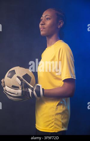 Femme afro-américaine debout dans un studio tenant un ballon de football, portant un maillot jaune et des gants Banque D'Images