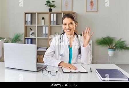 Médecin féminin sympathique regardant la caméra ayant un appel vidéo en ligne ou une conférence dans le bureau. Banque D'Images