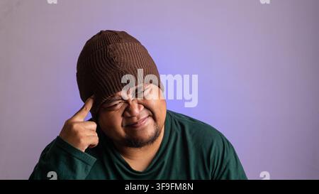 Un homme d'Asie du Sud-Est expressif touche la tempe d'un doigt tout en plissant les yeux dans une démonstration claire de tension mentale ou de profonde réflexion. Sur un fond Banque D'Images