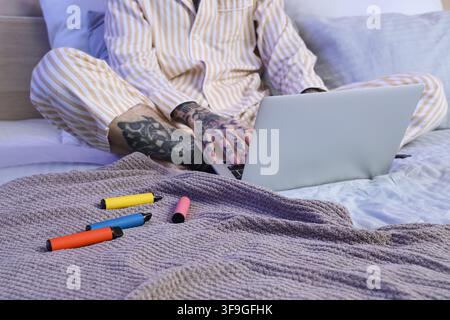 Jeune homme beau en pyjama avec différentes cigarettes électroniques jetables et ordinateur portable sur le lit à la maison Banque D'Images