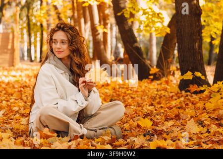 Jeune femme souriante assise dans le feuillage d'automne sur le sol de la forêt avec les feuilles jaunes Banque D'Images
