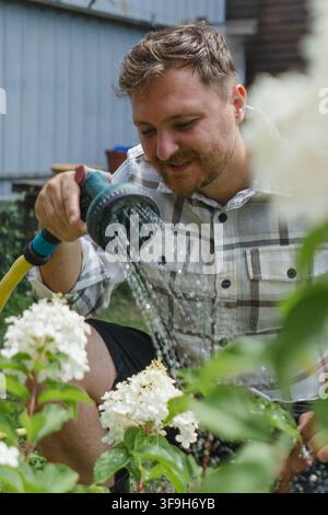 Jeune jardinier arrosant des arbustes d'hortensia blancs pendant l'été pour maintenir les plantes ornementales saines et florissantes dans un jardin domestique. Banque D'Images