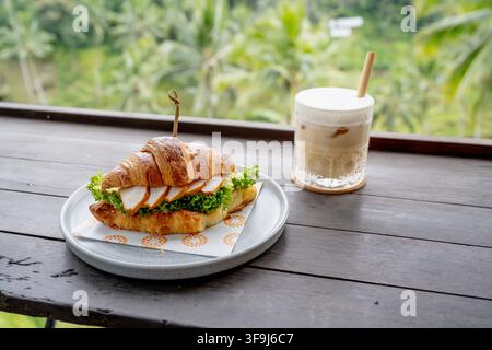 BALI, INDONÉSIE – 20 OCTOBRE 2024 : Sandwich gourmet au croissant et café servis sur un comptoir en bois donnant sur une verdure tropicale à Alas Harum. Banque D'Images