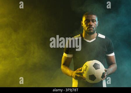 Homme africain tenant un ballon de football portant un maillot noir à bordures blanches dans une brume jaune bleu-vert en studio Banque D'Images