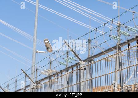 Une caméra de surveillance est montée sur un poteau près d'une clôture en fil de fer barbelé, avec des lignes électriques et des infrastructures de sous-station en arrière-plan Banque D'Images