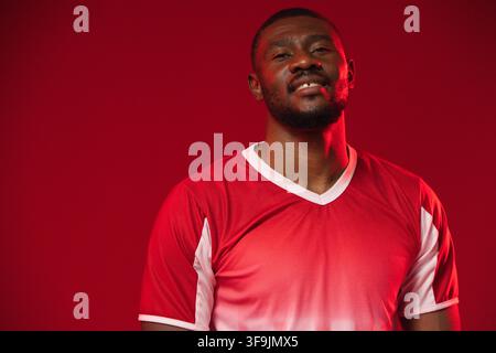 Homme d'âge moyen posant en studio avec un fond rouge, portant un maillot de sport rouge et une barbe courte Banque D'Images